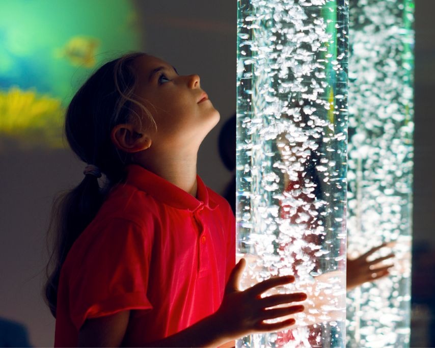 Young girl gazes up at a glowing bubble tube in an airport sensory room
