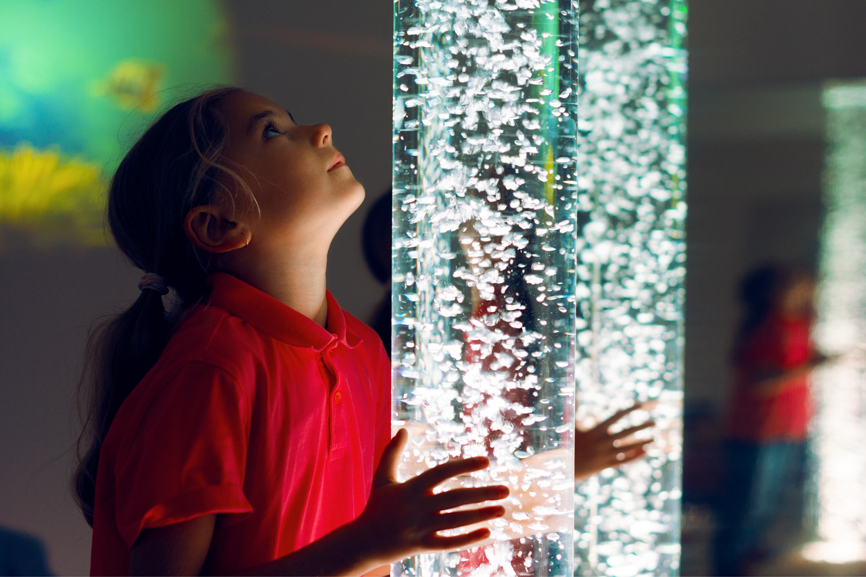 Young girl gazes up at a glowing bubble tube in an airport sensory room